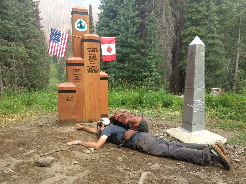 A Pacific Crest Trail thru-hiker struggling to touch the Northern PCT Terminus, marking the completion of the hike.