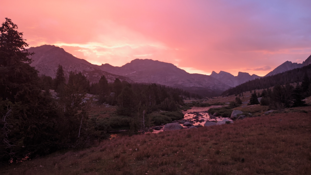 Sunrise glowing pink and orange over some mountain peaks. There is one substantially jagged peak. A stream runs through a valley, reflecting pinks and purples from the sunrise.
