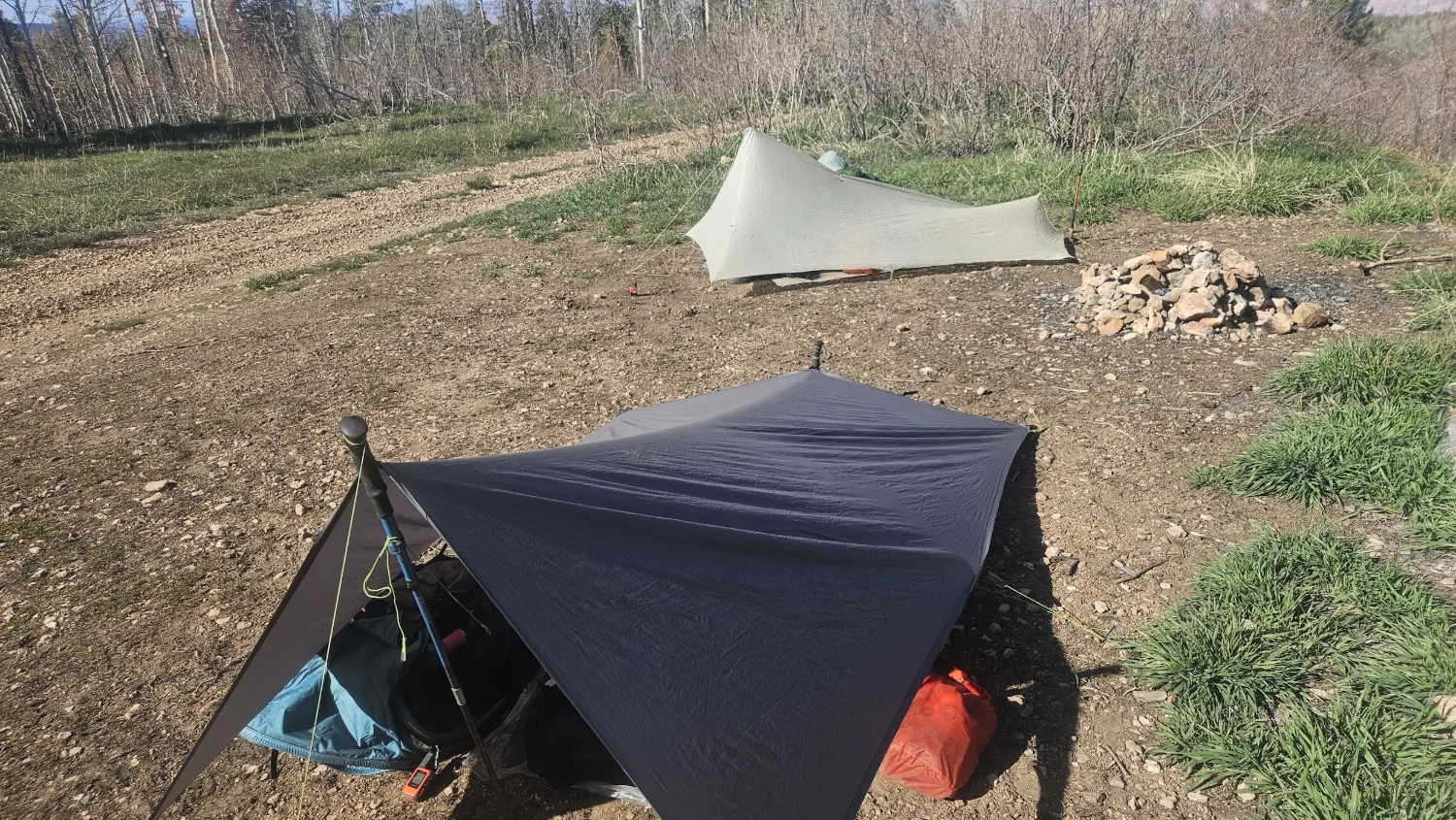 Low A-frame tarp pitch with far end staked close to the ground, backpack visible at the low end. A second tarp pitched higher in the background.