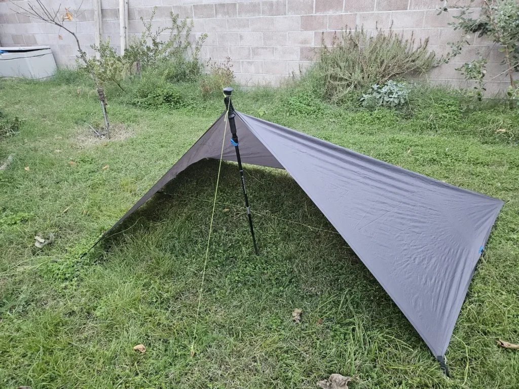 Half-pyramid tarp pitch in a backyard -- one end raised on a trekking pole, the other end staked flat to the ground